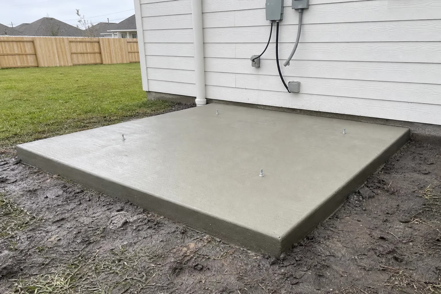 New concrete equipment pad with anchor bolts beside a house on a Houston-area lot